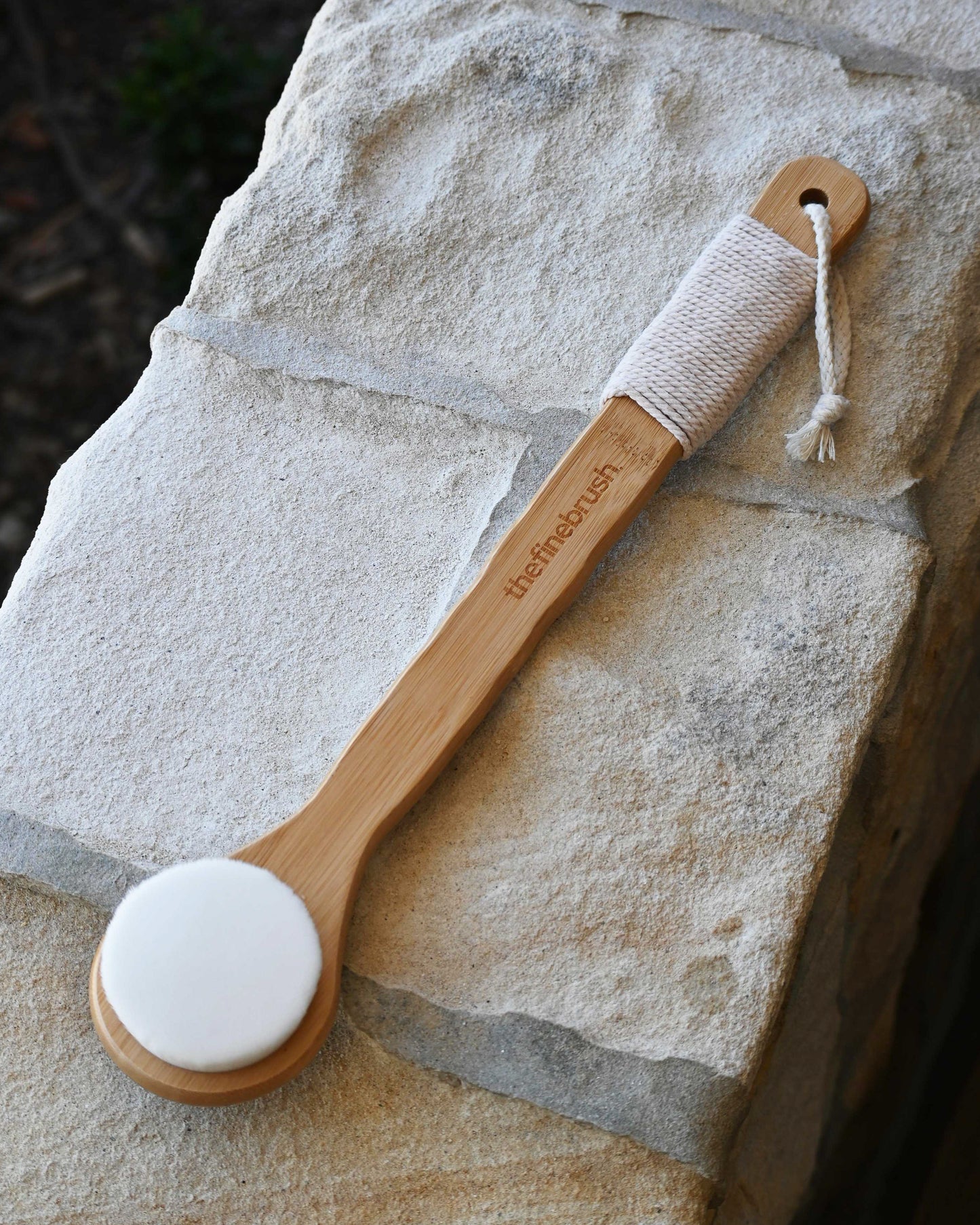 Full-length bamboo body brush with soft cotton loop, resting against stone — gentle enough for sensitive skin